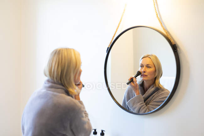 Glückliche ältere kaukasische Frau im Badezimmer, den Spiegel anschauend, sich schminkend. Lebensstil im Ruhestand, Zeit zu Hause verbringen. — Stockfoto