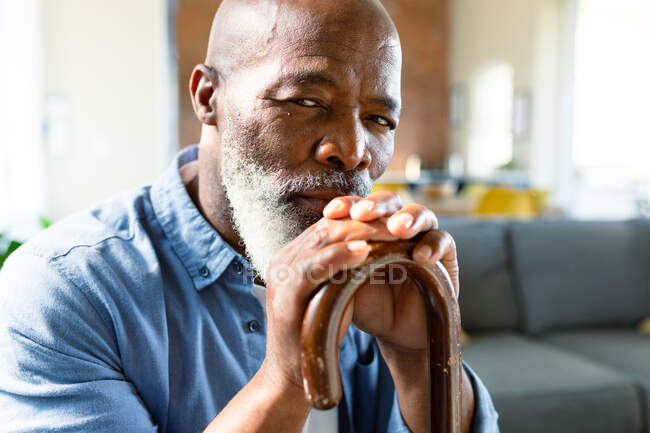 Retrato del reflexivo hombre afroamericano mayor en la sala de estar sosteniendo bastón. estilo de vida de jubilación, pasar tiempo en casa. - foto de stock