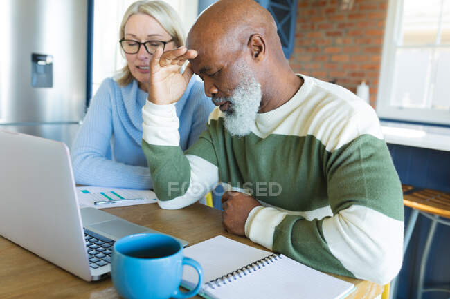 Stressed senior diverse couple in kitchen sitting at table, using laptop. retirement lifestyle, at home with technology. — Stock Photo