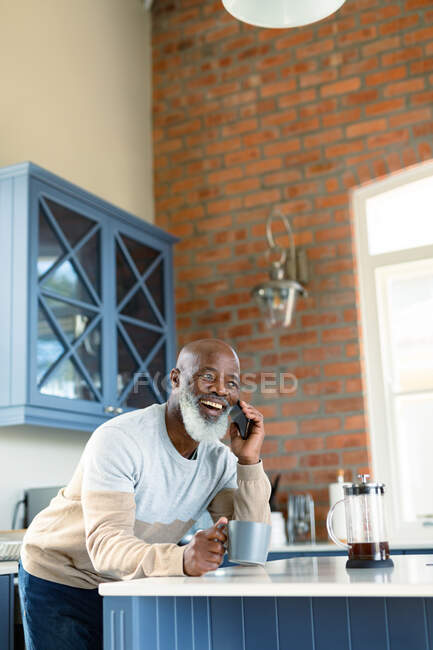 Glücklicher älterer afrikanisch-amerikanischer Mann in der Küche, der mit dem Smartphone spricht. Lebensstil im Ruhestand, zu Hause mit Technologie. — Stockfoto