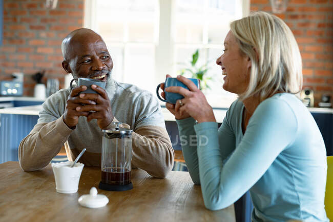 Feliz pareja de ancianos diversos en la cocina sentado en la mesa, tomando café. estilo de vida de jubilación, pasar tiempo en casa. - foto de stock