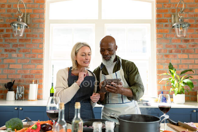 Felice coppia anziana diversificata in cucina indossando grembiuli, cucinare insieme, utilizzando tablet. stile di vita sano e attivo pensionamento a casa. — Foto stock