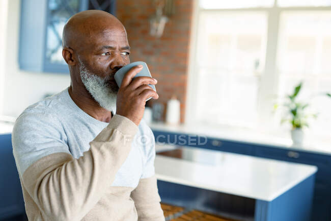 Pensativo hombre afroamericano mayor en la cocina bebiendo café, mirando hacia otro lado. estilo de vida de jubilación, pasar tiempo en casa. - foto de stock