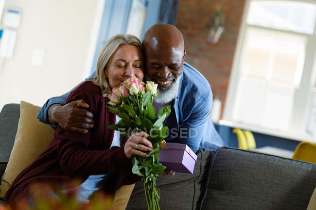 Щаслива старша різноманітна пара у вітальні сидить на дивані, даруючи квіти та подарунки. пенсійний спосіб життя, проводити час вдома . — стокове фото