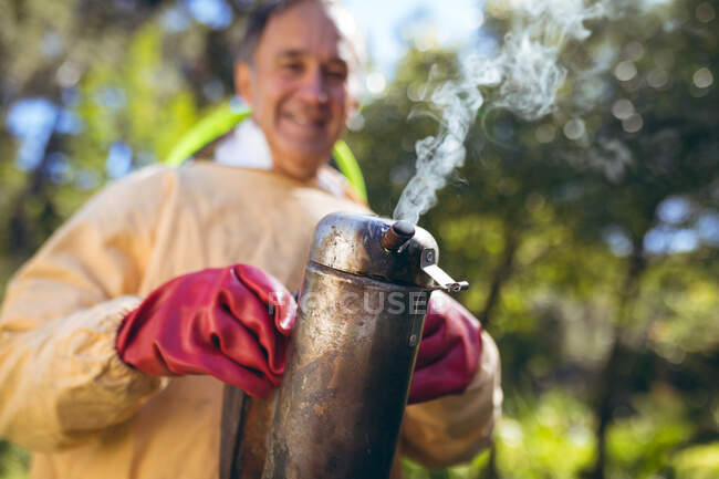Hombre mayor caucásico con uniforme de apicultor preparando humo para calmar a las abejas. apicultura, apicultura y producción de miel. - foto de stock