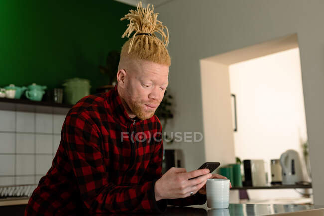 Nachdenklicher Albino-Afrikaner mit Dreadlocks, der Smartphone benutzt und Kaffee trinkt. Fernbedienung mit Technologie zu Hause. — Stockfoto
