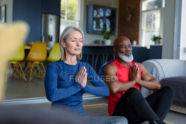 Happy senior diverse couple in exercise clothes practicing yoga together, meditating. healthy, active retirement lifestyle at home. — Stock Photo