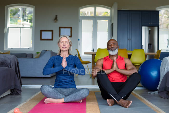 Feliz pareja mayor diversa en ropa de ejercicio practicando yoga juntos, meditando. estilo de vida saludable y activo de jubilación en el hogar. - foto de stock