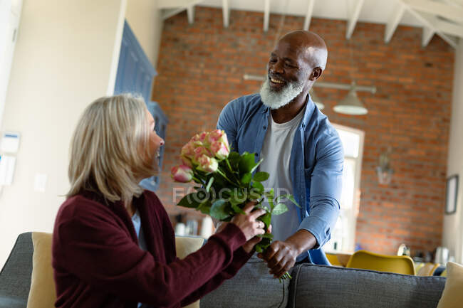 Щаслива старша різноманітна пара у вітальні сидить на дивані, даруючи квіти. пенсійний спосіб життя, проводити час вдома . — стокове фото