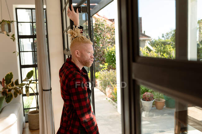 Pensativo albino hombre afroamericano con rastas mirando por la ventana. trabajo remoto utilizando tecnología en el hogar. - foto de stock