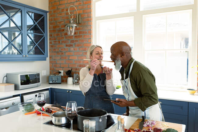 Happy senior diverse couple in kitchen wearing aprons, cooking together, trying food. healthy, active retirement lifestyle at home. — Stock Photo