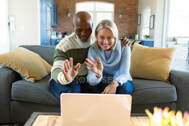 Щаслива старша різноманітна пара у вітальні сидить на дивані, використовуючи ноутбук, роблячи відеодзвінки. пенсійний спосіб життя, вдома з технологіями . — стокове фото