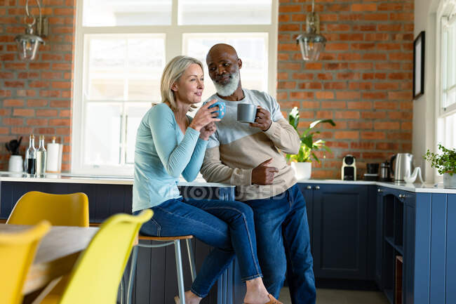 Feliz pareja de ancianos diversos en la cocina sentado en la encimera, bebiendo café. estilo de vida de jubilación, pasar tiempo en casa. - foto de stock
