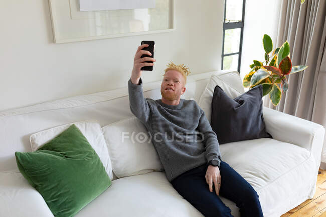 Homem americano africano albino na sala de estar a tirar selfie. tempo de lazer usando a tecnologia, relaxando em casa. — Fotografia de Stock