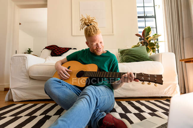 Улыбающийся американец-альбинос, сидящий на полу в гостиной и играющий на гитаре. свободное время, отдых дома. — стоковое фото