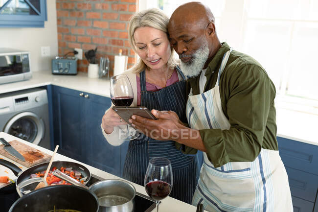 Feliz casal diversificado sênior na cozinha vestindo aventais, cozinhando juntos, usando tablet. estilo de vida saudável e ativo em casa. — Fotografia de Stock
