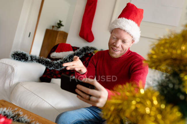 Hombre afroamericano albino con sombrero de santa haciendo videollamada con decoraciones navideñas. navidad, fiesta y tecnología de la comunicación festividad y tecnología de la comunicación. - foto de stock