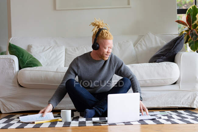 Альбинос африканский американец, сидящий на полу и работающий из дома на ноутбуке. удаленная работа с использованием технологии на дому. — стоковое фото