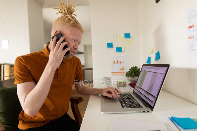 Hombre afroamericano albino trabajando desde casa y haciendo una llamada telefónica y usando un portátil. trabajo remoto utilizando tecnología en el hogar. - foto de stock