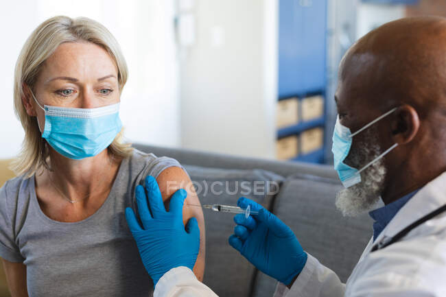 Happy senior diverse woman and doctor wearing face masks in living room sitting on sofa, vaccinating. senior health and lifestyle during covid 19 pandemic. — Stock Photo