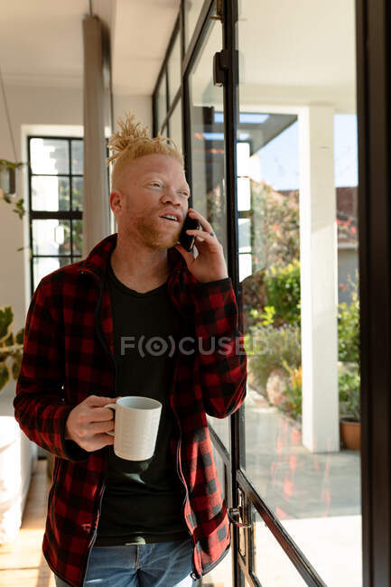 Lächelnder Albino-Afrikaner mit Dreadlocks, der Smartphone benutzt und Kaffee trinkt. Fernbedienung mit Technologie zu Hause. — Stockfoto