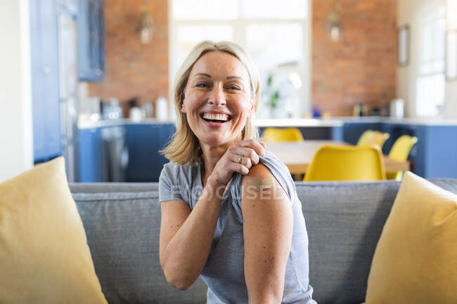 Feliz mujer caucásica mayor en la sala de estar con vendaje en el brazo. salud y estilo de vida durante la pandemia de covid 19. - foto de stock