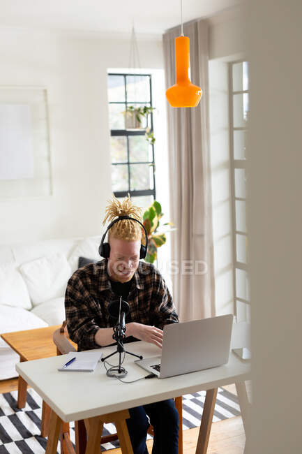 Feliz albino hombre afroamericano con rastas trabajando desde casa y haciendo podcast. trabajo remoto utilizando tecnología en el hogar. - foto de stock