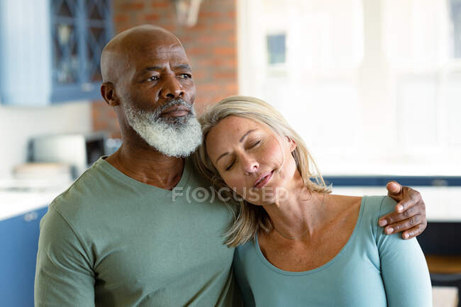 Feliz pareja de ancianos diversos en la cocina abrazando con los ojos cerrados. estilo de vida de jubilación, pasar tiempo en casa. - foto de stock