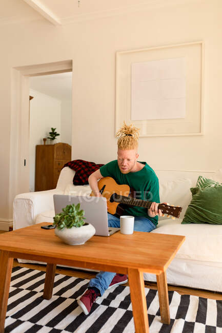 Albino homem americano africano na sala de estar tocando guitarra e usando laptop. tempo de lazer usando a tecnologia, relaxando em casa. — Fotografia de Stock