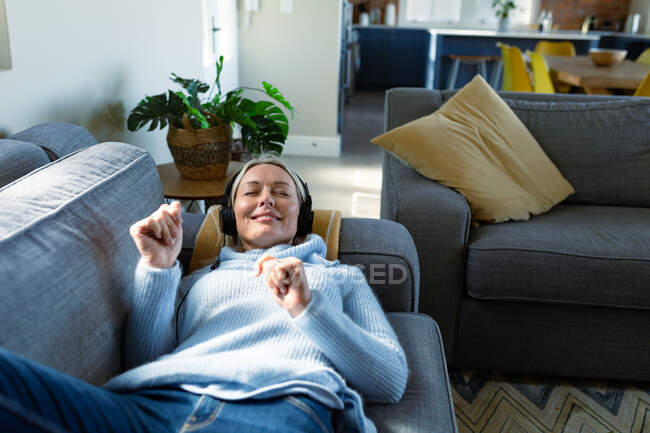 Щаслива старша біла жінка у вітальні лежить на дивані, одягнена в навушники. пенсійний спосіб життя, вдома з технологіями . — стокове фото