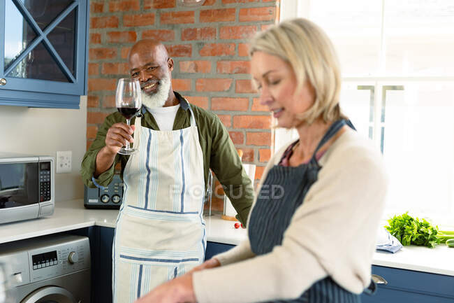 Feliz casal diversificado sênior na cozinha vestindo aventais, cozinhando juntos, bebendo vinho. estilo de vida saudável e ativo em casa. — Fotografia de Stock