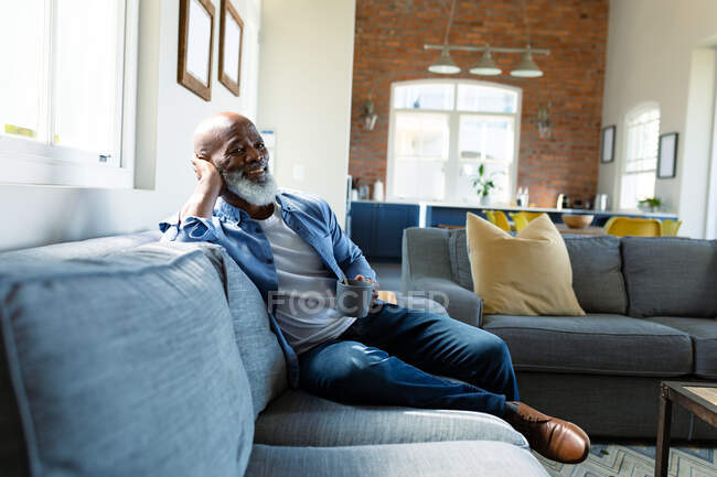 Heureux homme afro-américain senior dans le salon assis sur le canapé, tenant une tasse. mode de vie à la retraite, passer du temps chez soi. — Photo de stock