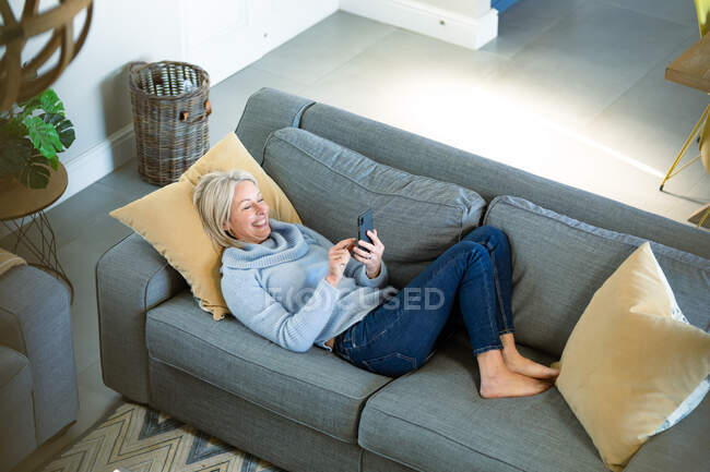 Feliz anciano mujer caucásica en la sala de estar tumbado en el sofá, utilizando el teléfono inteligente. estilo de vida de jubilación, en casa con tecnología. - foto de stock
