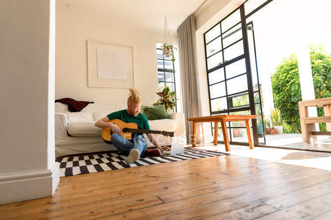 Albino-amerikanischer Mann im Wohnzimmer, der Gitarre spielt und Laptop benutzt. Freizeit mit Technik, Entspannung zu Hause. — Stockfoto