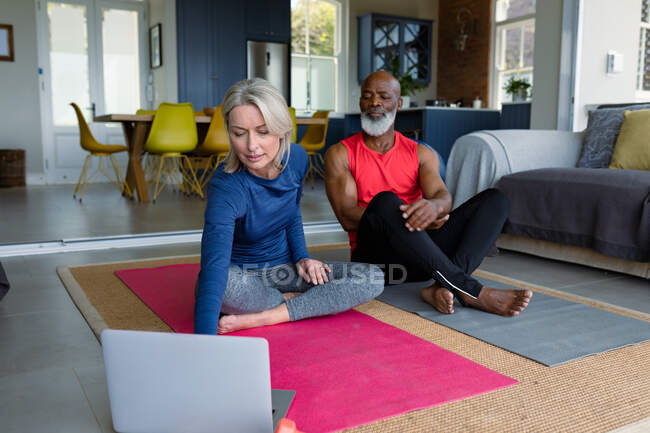Felice coppia anziana diversificata in abiti da ginnastica praticare yoga insieme, utilizzando il computer portatile. stile di vita sano e attivo pensionamento a casa. — Foto stock