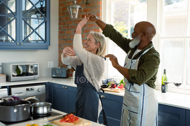 Happy senior diverse couple in kitchen wearing aprons, cooking together, dancing. healthy, active retirement lifestyle at home. — Stock Photo