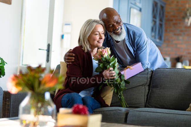 Feliz pareja de ancianos diversos en la sala de estar sentado en el sofá, dando flores y regalos. estilo de vida de jubilación, pasar tiempo en casa. - foto de stock