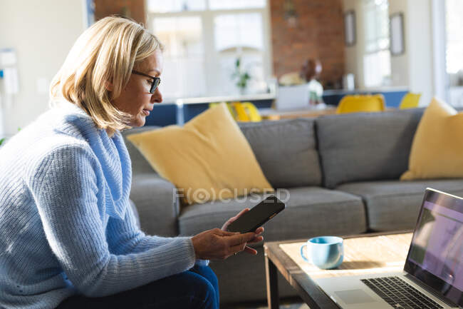 Feliz anciano mujer caucásica en la sala de estar sentado en el sofá, utilizando el teléfono inteligente y el ordenador portátil. estilo de vida de jubilación, en casa con tecnología. - foto de stock
