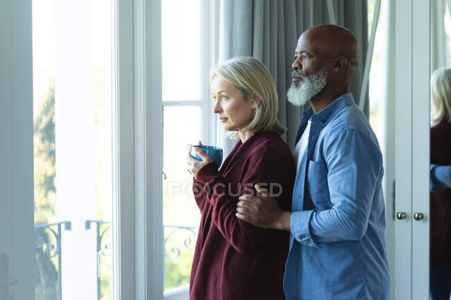 Besorgte Senioren blicken durchs Fenster und umarmen sich. Lebensstil im Ruhestand, Zeit zu Hause verbringen. — Stockfoto