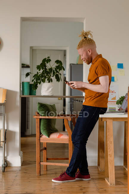Homme souriant albinos afro-américain avec dreadlocks à l'aide d'un smartphone et boire du café. télétravail utilisant la technologie à la maison. — Photo de stock