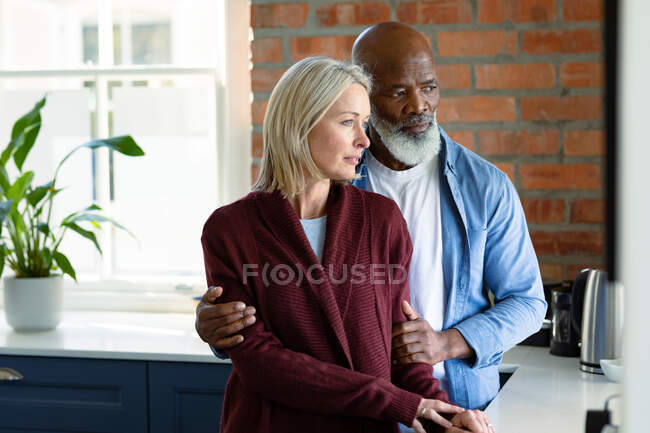 Preoccupato anziano coppia diversificata in cucina appoggiata sul piano di lavoro, abbracciando e guardando altrove. stile di vita di pensione, trascorrere del tempo a casa. — Foto stock