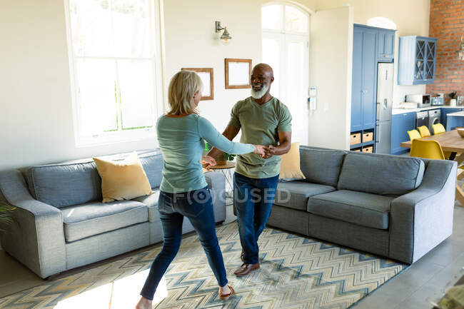 Feliz casal diverso sênior na sala de estar dançando juntos. estilo de vida da aposentadoria, passar tempo em casa. — Fotografia de Stock