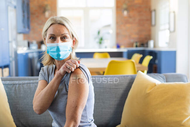 Mulher caucasiana sênior feliz usando máscara facial na sala de estar com bandagem no braço. saúde sênior e estilo de vida durante a pandemia covid 19. — Fotografia de Stock