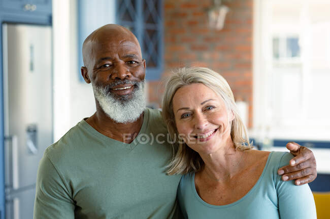 Portrait de heureux couple de personnes âgées diversifiées dans la cuisine embrassant et souriant. mode de vie à la retraite, passer du temps chez soi. — Photo de stock