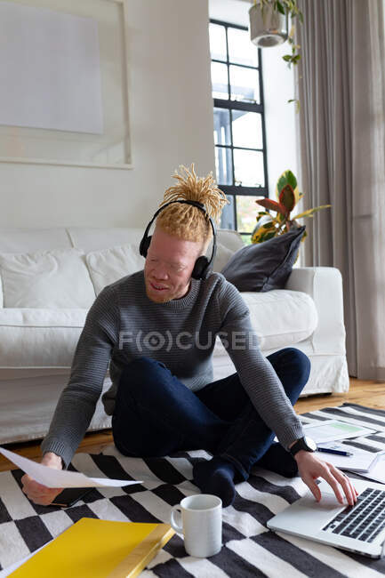 Альбинос африканский американец, сидящий на полу и работающий из дома на ноутбуке. удаленная работа с использованием технологии на дому. — стоковое фото