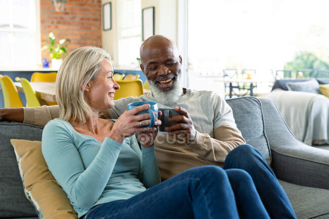 Feliz pareja de ancianos diversos en la sala de estar sentado en el sofá, beber café. estilo de vida de jubilación, pasar tiempo en casa. - foto de stock