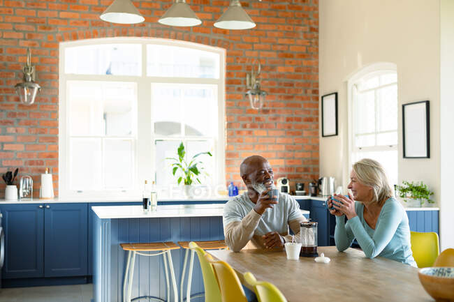 Happy senior diverse couple in kitchen sitting at table, drinking coffee. retirement lifestyle, spending time at home. — Stock Photo