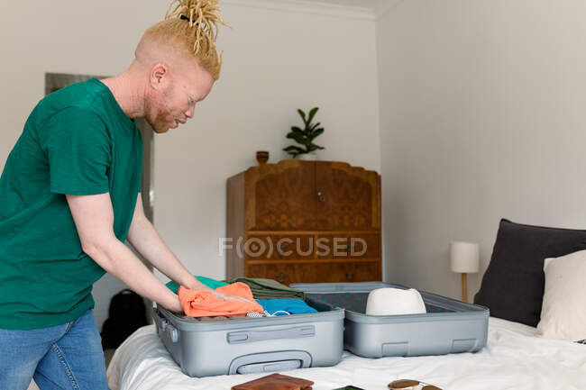 Albino afroamericano hombre empacando maleta en el dormitorio. preparación de vacaciones y viajes durante la pandemia de covid 19. - foto de stock