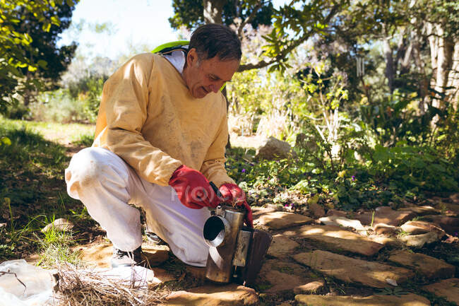 Caucasian senior man wearing beekeeper uniform preparing smoke to calm bees. beekeeping, apiary and honey production concept. — Stock Photo
