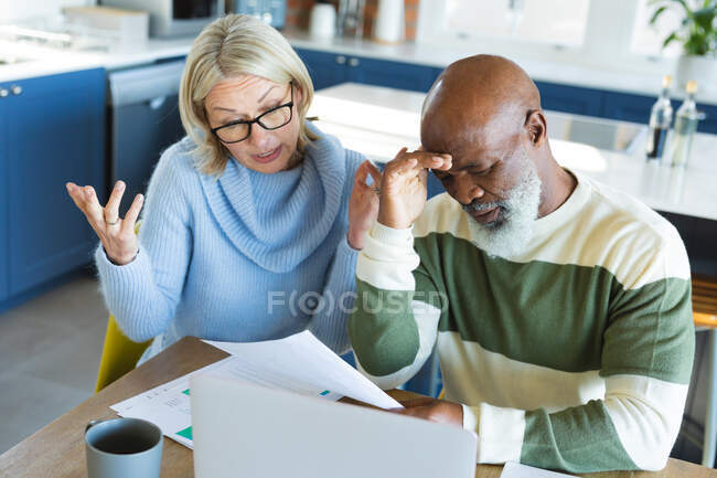 Sforzato anziani coppia diversificata in cucina seduto a tavola, utilizzando il computer portatile. stile di vita di pensione, a casa con la tecnologia. — Foto stock
